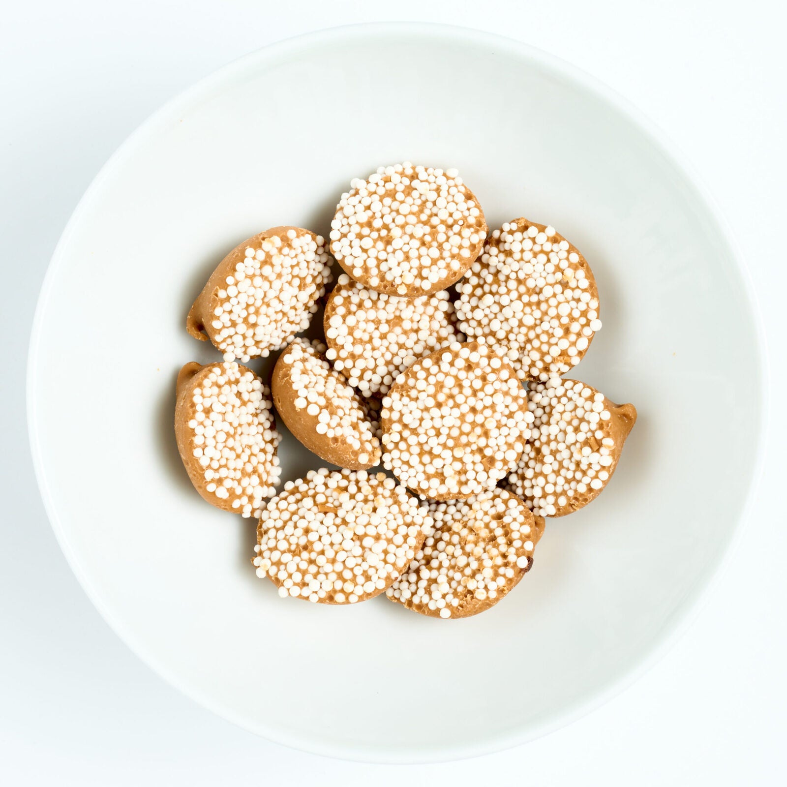 Peanut butter nonpareils topped with tiny white sprinkles, round candies arranged on a white plate.
