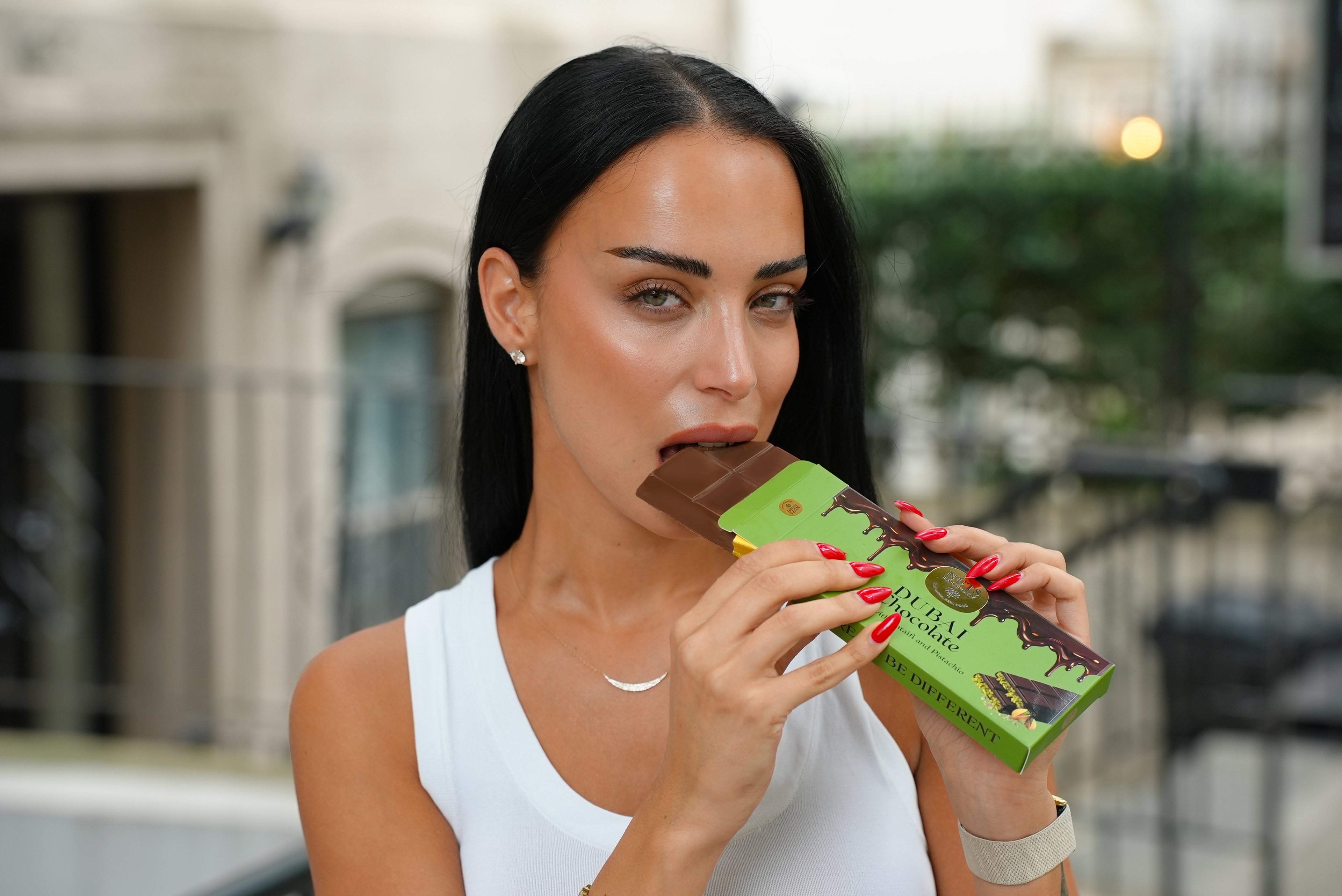 A woman with long black hair and red nails is shown in a lifestyle setting, eating a chocolate bar wrapped in green packaging that reads 'Dubai Chocolate'.